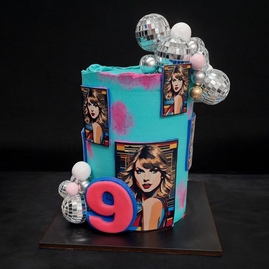 This is a vibrant, Taylor Swift-themed birthday cake with a bold, artistic design. The cake features a tall, cylindrical shape with bright turquoise buttercream and pink airbrushed accents. It is decorated with multiple pop-art style edible images of Taylor Swift, giving it a modern and stylish look. A large fondant number "9" in pink with a blue outline is placed on the front, signifying the birthday celebration. The cake is also adorned with clusters of silver disco balls, metallic edible pearls, and whit