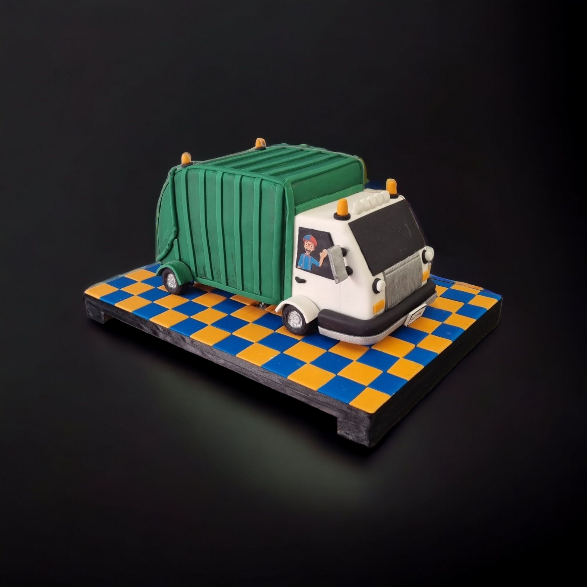 "Blippi-themed dump truck cake designed with vibrant green and white fondant, featuring a truck driver detail, displayed on a colorful blue and orange checkered base against a black background."