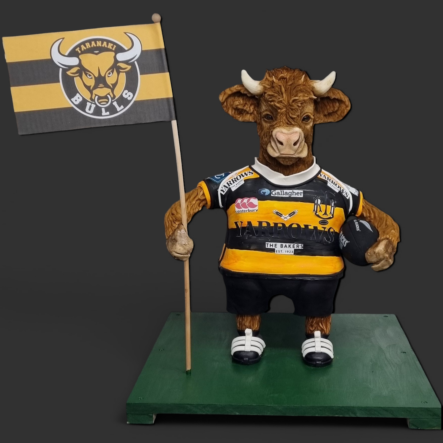 "Taranaki Bulls-themed sculpted cake featuring a detailed highlander bull mascot wearing a rugby jersey, holding a team flag and rugby ball, standing on a green base with a gray background."