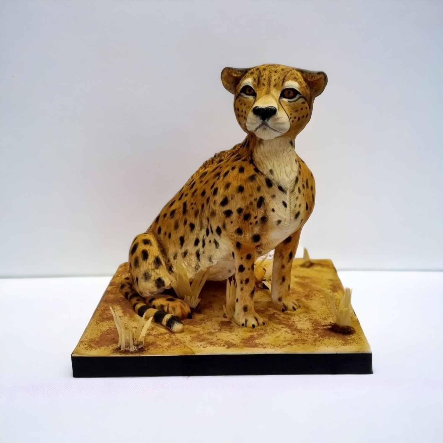 Realistic sculpted cheetah cake featuring detailed spots and lifelike textures, seated on a savanna-inspired base with dry grass accents, displayed against a white background.