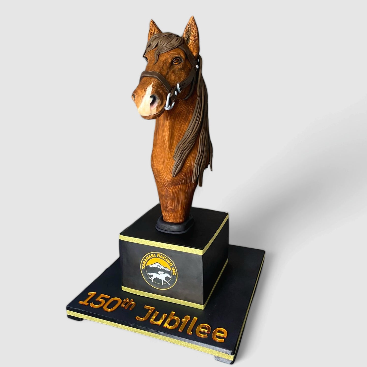 "Elegant horse head sculpted cake created for the 150th Jubilee of Taranaki Racing, featuring intricate detailing, a bridle, and a custom logo base with gold lettering, displayed on a clean white background.