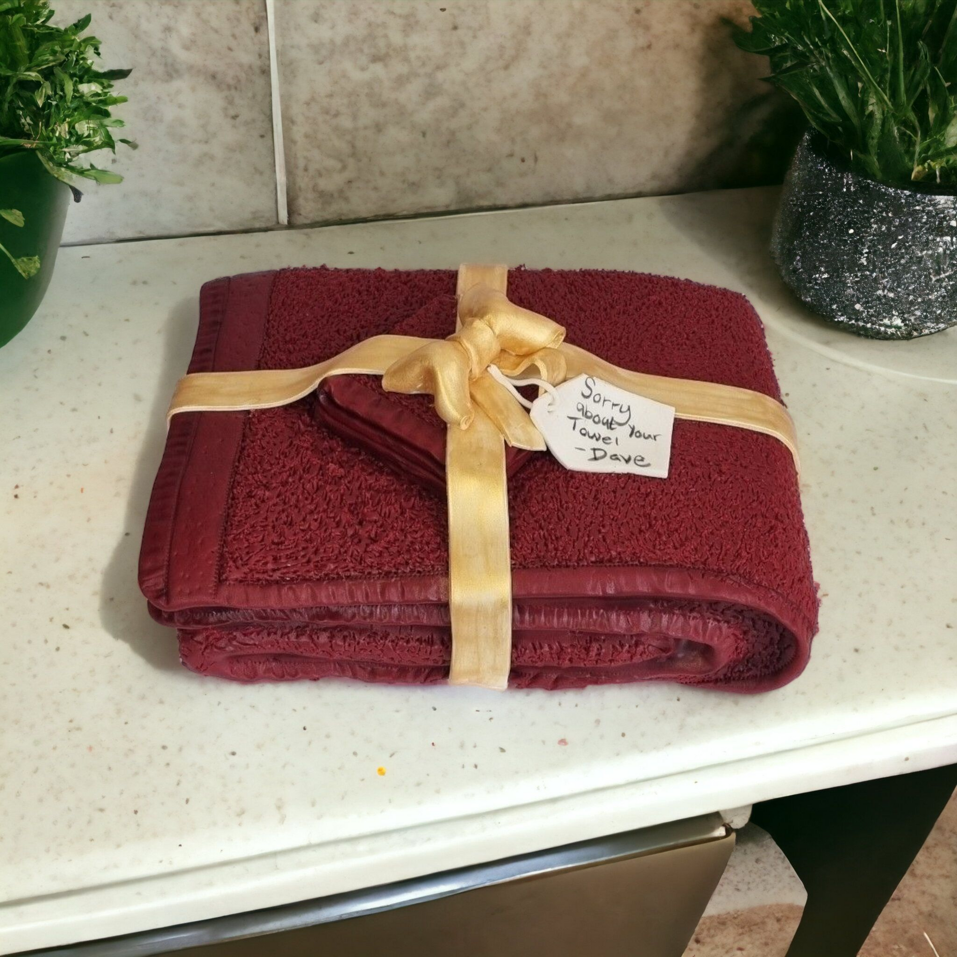 "Realistic sculpted cake designed to look like a folded maroon towel, wrapped with a golden ribbon and a handwritten tag reading 'Sorry about your towel - Dave,' displayed on a bathroom counter with decorative plants.