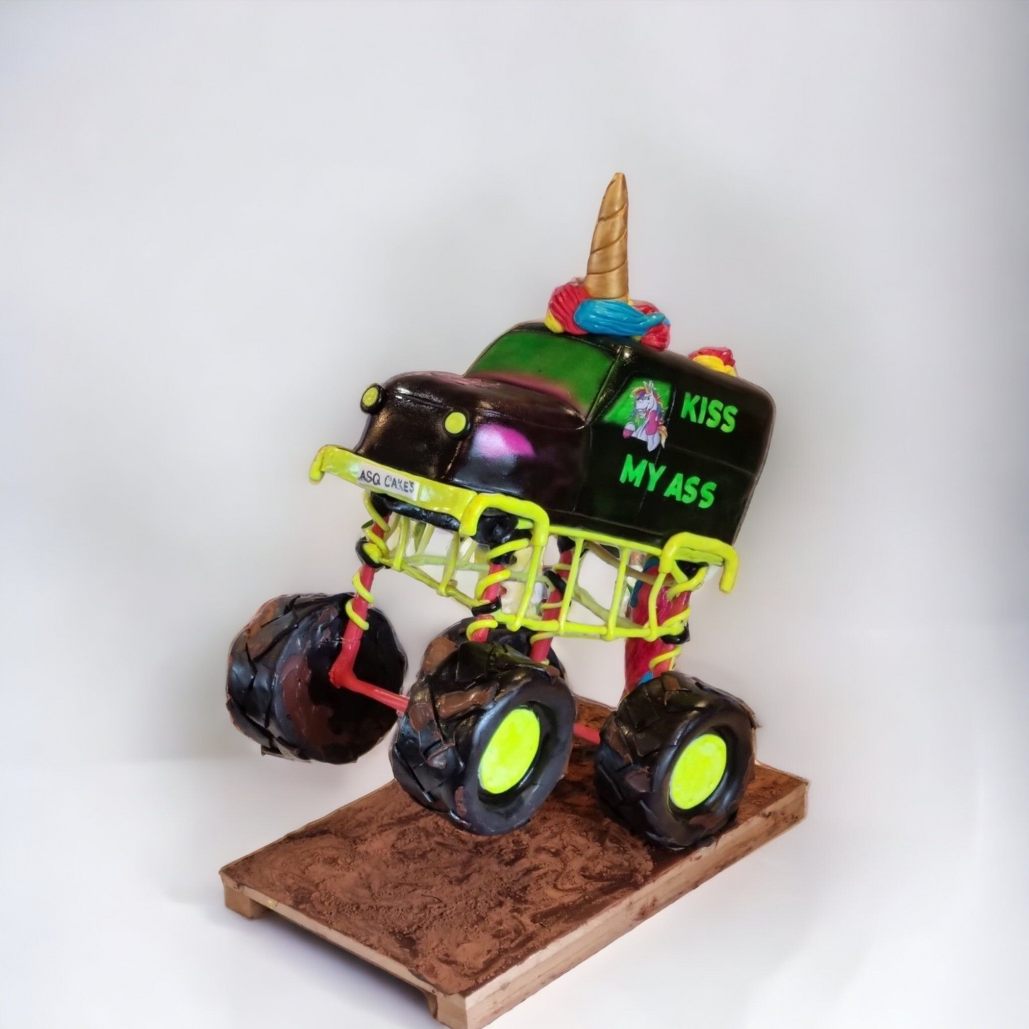 "Unicorn-themed monster truck cake performing a wheelie, featuring vibrant green and black design, a golden horn, and playful 'Kiss My Ass' graphics, displayed on a textured brown base against a light background."